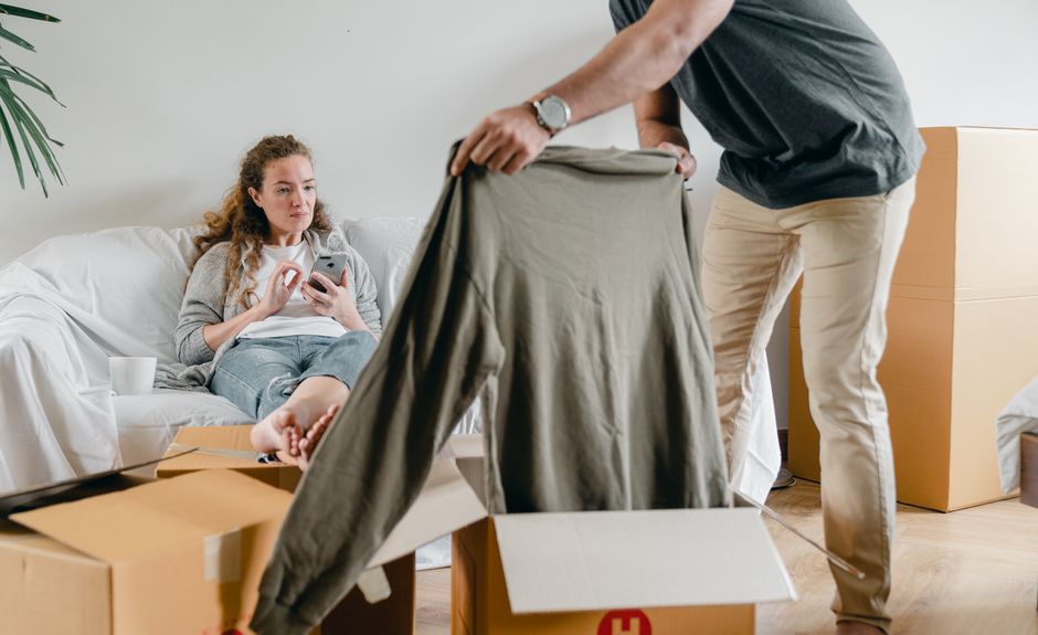 Crop anonymous male in watch getting polo neck out of cardboard box while standing near pensive barefoot girlfriend sitting with crossed legs on comfortable couch after moving into new flat